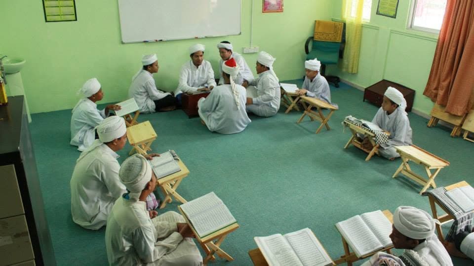 Pelajar duduk dalam bulatan di rehal masing-masing dengan mushaf terbuka, dibimbing asatizah di tengah bilik darjah